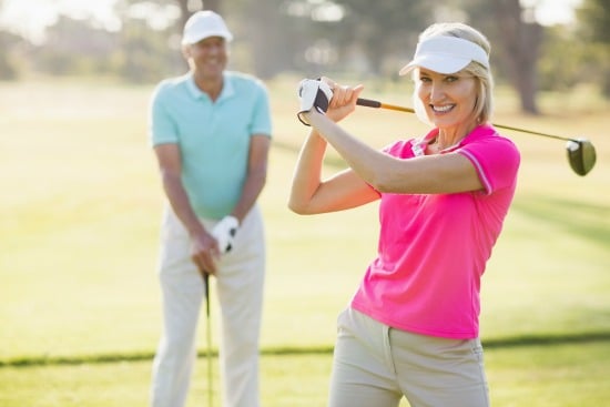senior woman in pink shirt smiling ad lifting a golf club while senior man looks on in background